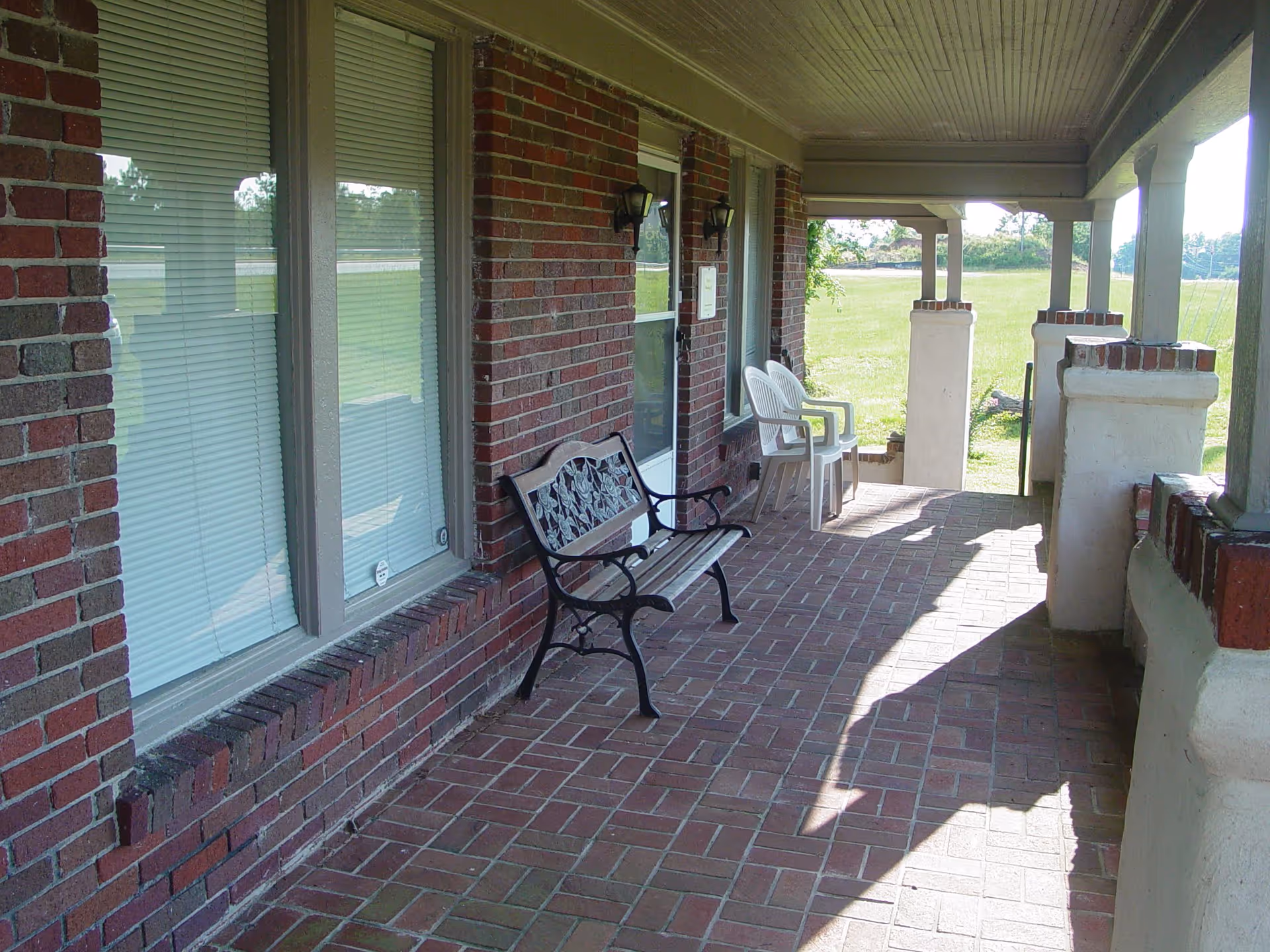 Brick covered front porch with a metal bench and plastic chairs overlooking a grassy yard.