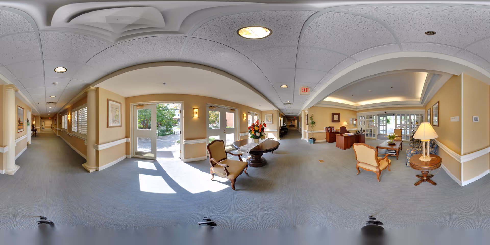 A spacious and well-lit senior living facility interior with beige walls and blue carpet. The area features a seating arrangement with classic wooden chairs and a round table with a flower arrangement. Large windows and glass doors allow natural light to enter, showing greenery outside. There is a reception desk with a lamp and additional seating in the background, and a long hallway extends to the left side of the image.