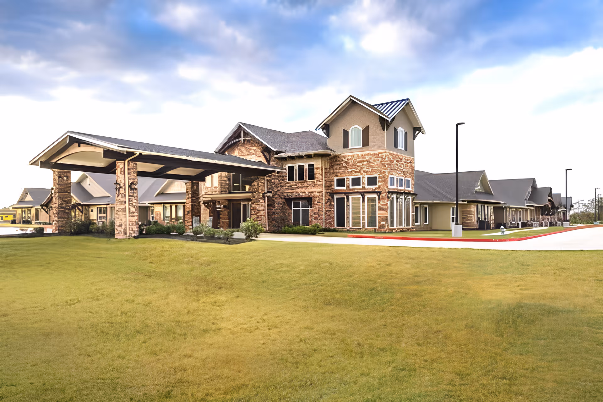 Exterior view of a large senior living facility building with stone and brick facade, multiple windows, and a covered entrance driveway. The building is surrounded by a well-maintained grassy area and a paved driveway with street lamps under a partly cloudy sky.