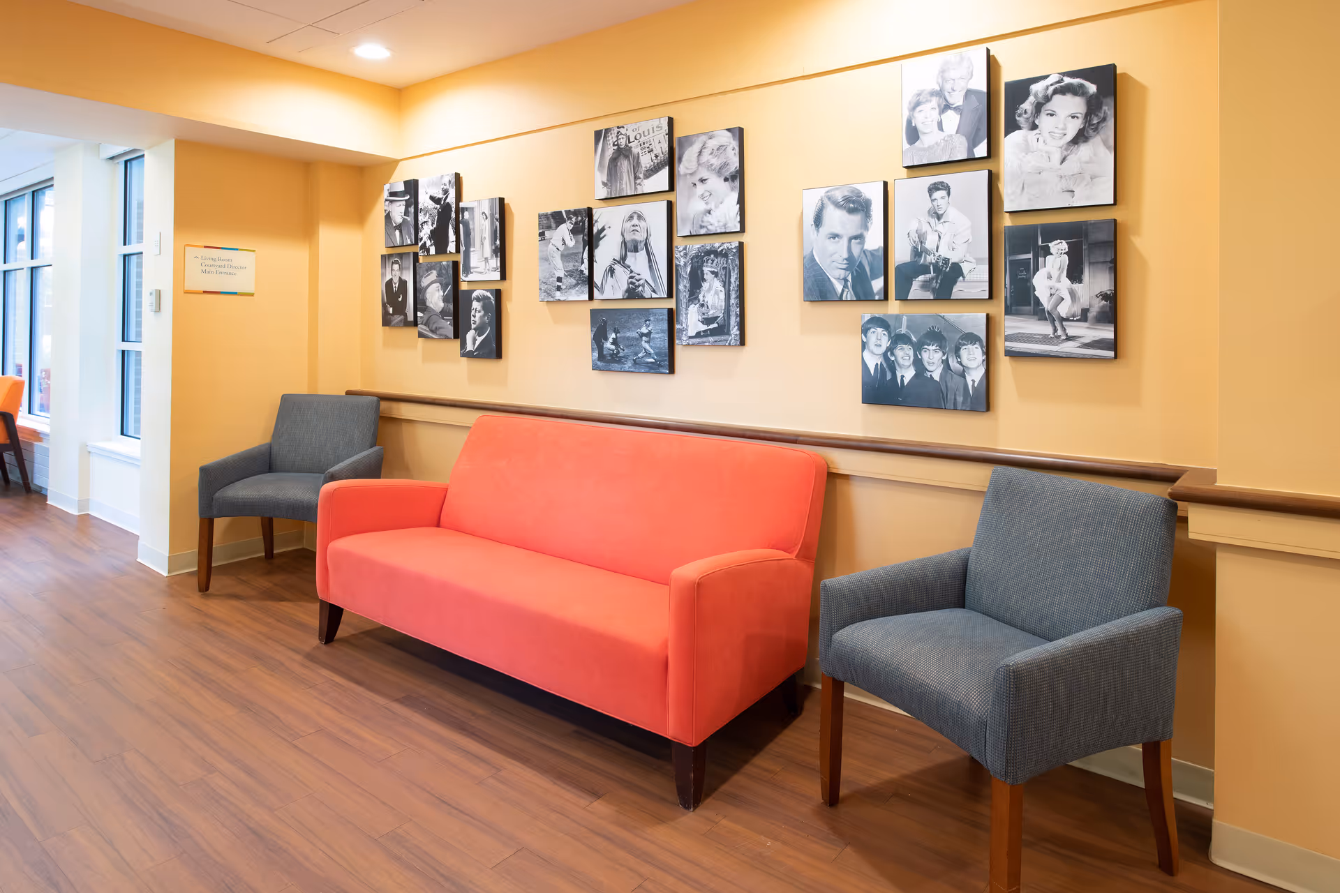 A seating area in a senior living facility with a bright orange couch flanked by two gray armchairs. The walls are painted a warm yellow and decorated with black and white photographs of famous personalities and scenes. The floor is wooden, and there is a window letting in natural light on the left side.