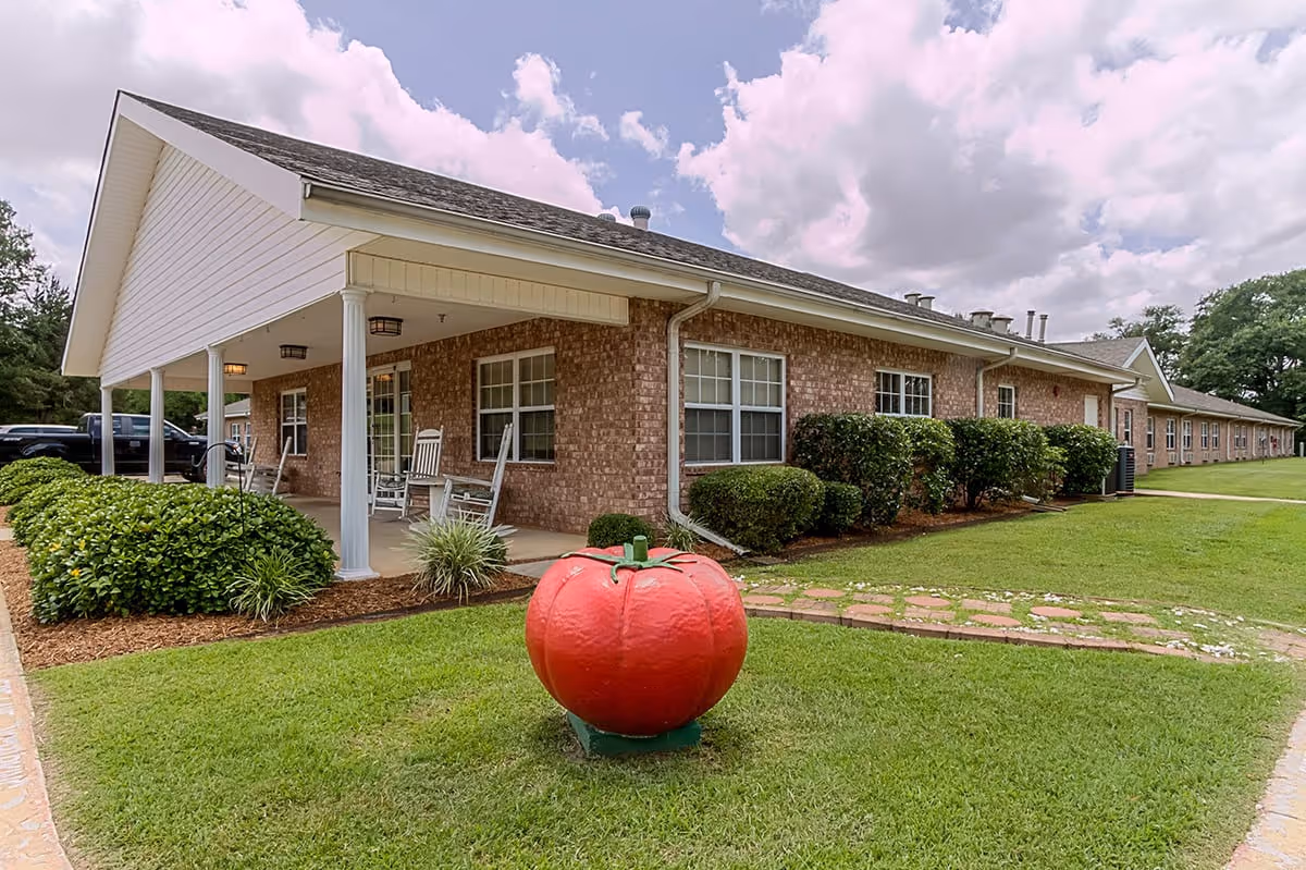 Exterior view of a single-story brick building with a covered porch featuring rocking chairs. The building is surrounded by green grass, bushes, and a large decorative red tomato sculpture in the foreground. The sky is partly cloudy.