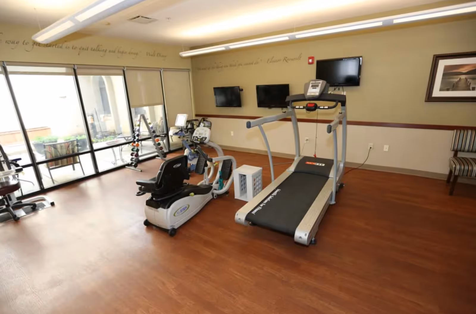 A fitness room with exercise equipment including a recumbent bike, a treadmill, and a stationary bike. The room has wooden flooring, large windows with blinds, two wall-mounted TVs, and a rack of dumbbells. There is a framed picture on the wall and a striped chair in the corner.