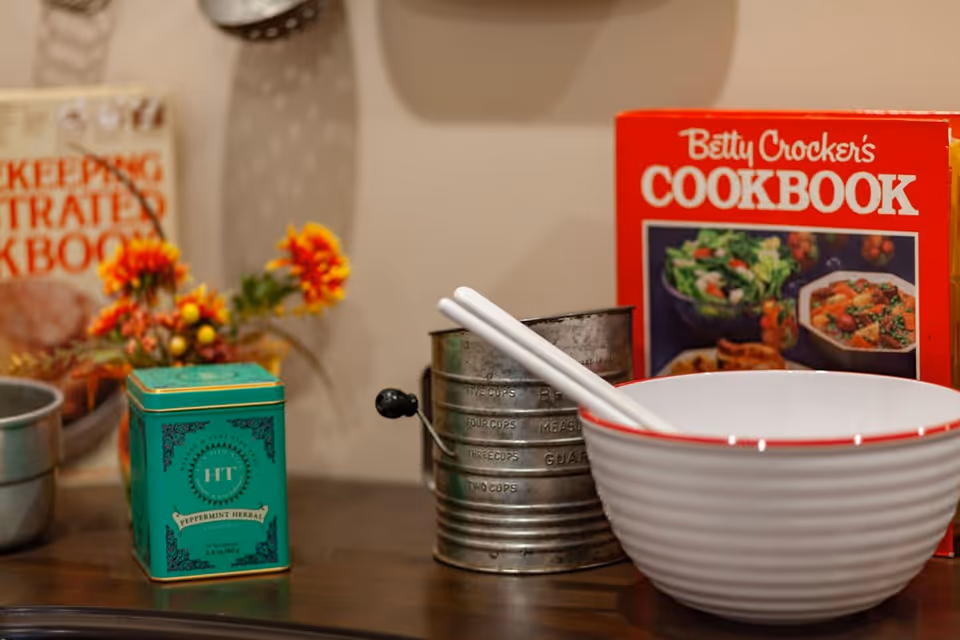 Close-up of a kitchen counter with a white bowl containing two white chopsticks, a vintage metal sifter, a green tin labeled 'Peppermint Herbal', a cookbook titled 'Betty Crocker's Cookbook' with images of food on the cover, and a small bouquet of orange and yellow flowers in the background.