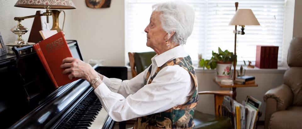 An elderly woman with white hair is sitting at a black piano, placing a red book titled 'PRAISE' on the music stand. The room is well-lit with natural light coming through a window with white blinds. There is a green chair, a side table with a potted plant, a lamp, and some books and magazines in the background.