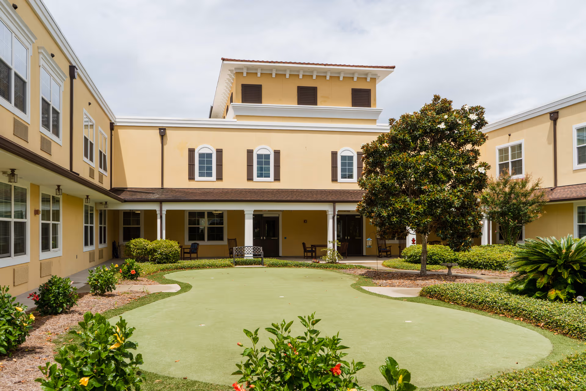 Outdoor courtyard area at The Beacon at Gulf Breeze featuring a putting green surrounded by landscaped bushes and flowering plants. The courtyard is enclosed by a two-story yellow building with white trim and brown shutters. There are several chairs and benches under a covered walkway around the courtyard.