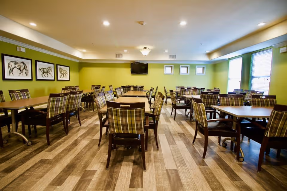 A dining room with multiple tables and chairs arranged neatly. The chairs have plaid upholstery and dark wooden frames. The walls are painted green and decorated with framed horse artwork. There is a flat-screen TV mounted on the far wall and windows with blinds letting in natural light.