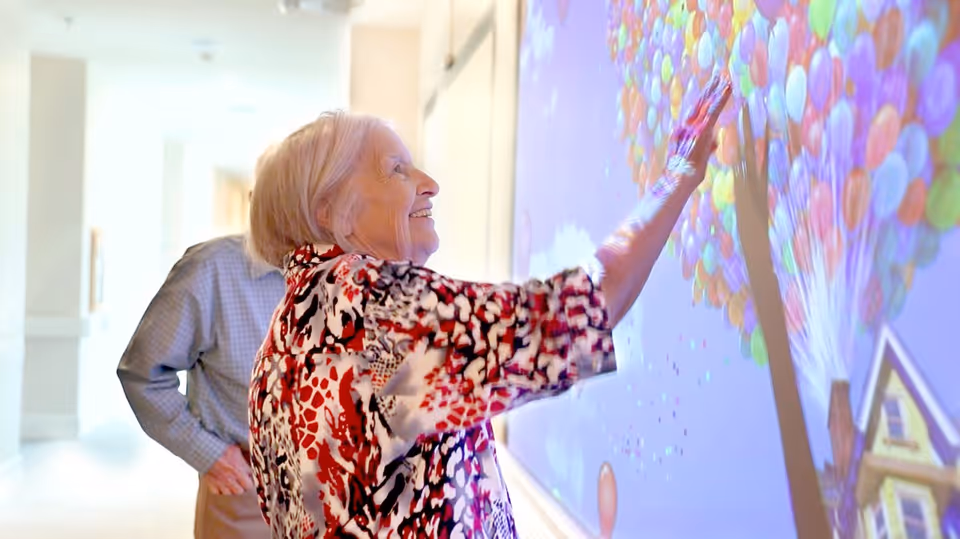 An elderly woman smiling and interacting with a colorful wall display featuring numerous balloons. A man stands behind her, watching. They are in a well-lit hallway with light-colored walls.