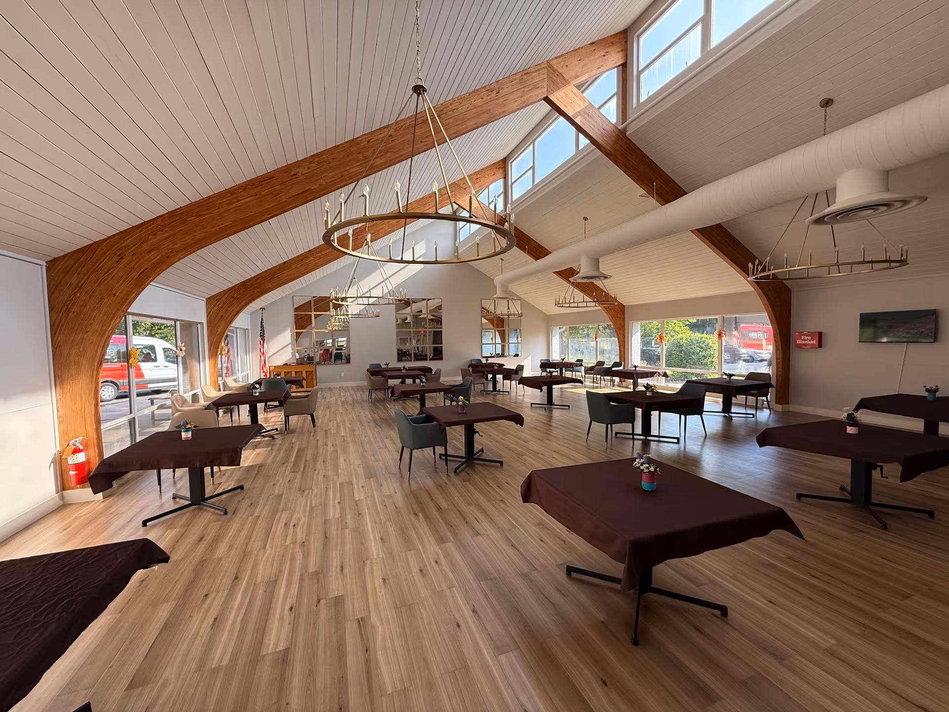 A spacious dining room with wooden beams on the ceiling and large windows letting in natural light. The room has multiple tables covered with dark brown tablecloths, each with a small flower arrangement. There are comfortable chairs around the tables and chandeliers hanging from the ceiling. The floor is light wood, and there is a fire extinguisher and a fire alarm on the walls.