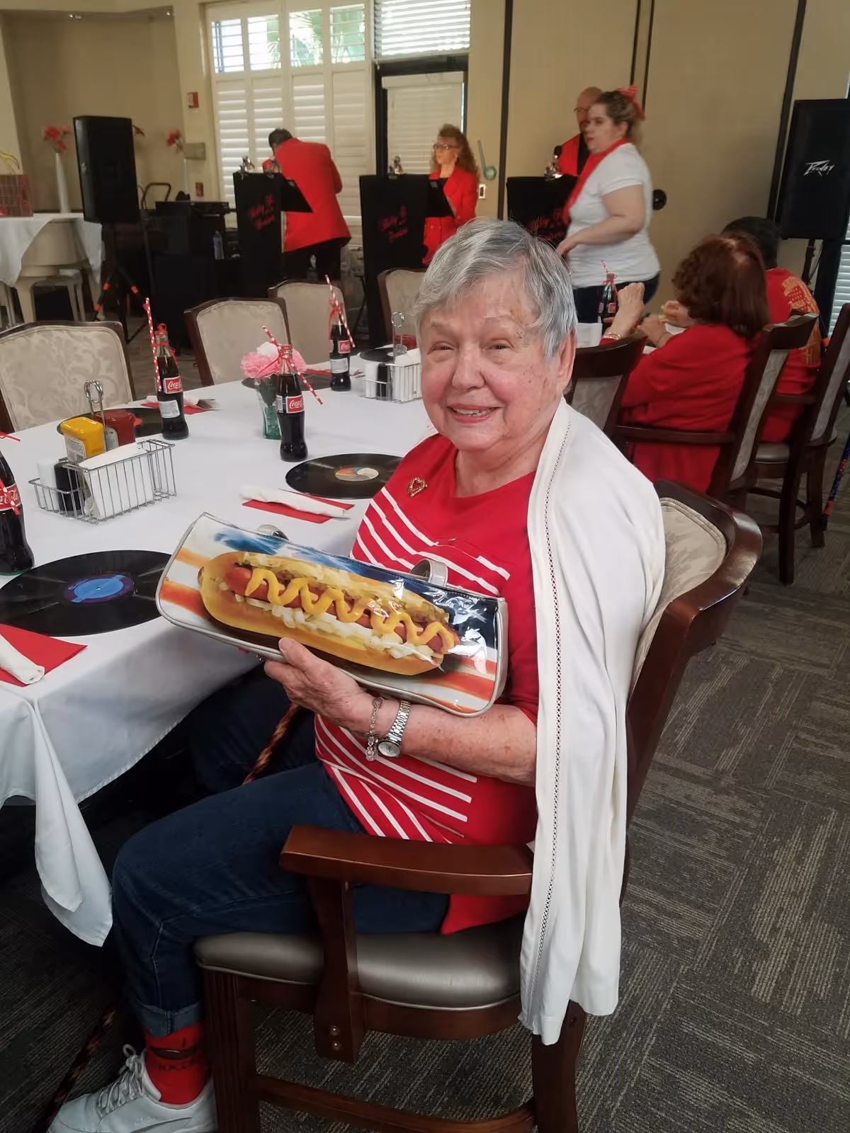 An elderly woman in a red-striped shirt smiles while seated at a decorated dining table holding a hot dog-themed clutch, with other people and decorations in the background.