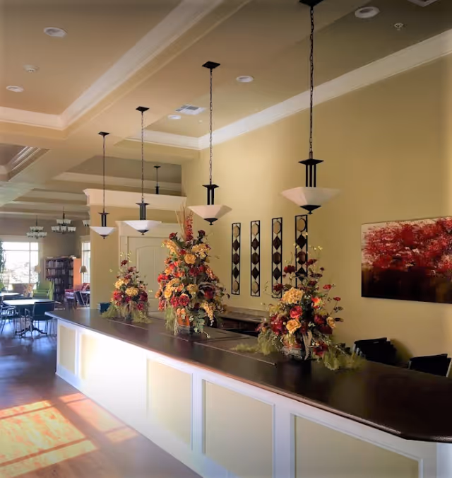 Interior view of a reception or common area in a retirement center featuring a long counter with three large floral arrangements on top. Above the counter, there are four hanging pendant lights. The walls are painted beige and decorated with a red and black abstract painting and four vertical decorative wall pieces. In the background, there are tables and chairs near large windows letting in natural light.