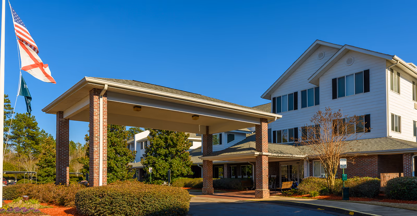 Exterior view of Monarch Estates senior living facility showing a covered entrance supported by brick columns, a three-story building with white siding and black shutters, surrounded by landscaped bushes and trees under a clear blue sky. Three flags are flying on a flagpole to the left of the entrance.