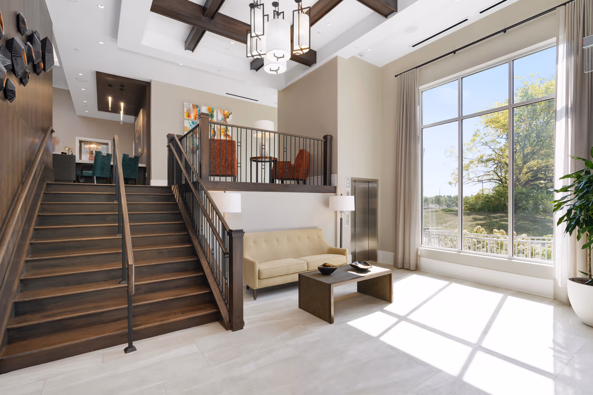 Bright and spacious interior of a senior living facility with a large window letting in natural light. The room features a beige sofa, a modern coffee table, a floor lamp, and a staircase with dark wooden railings leading to an upper seating area with orange chairs and a small table. The ceiling has exposed wooden beams and a contemporary chandelier.