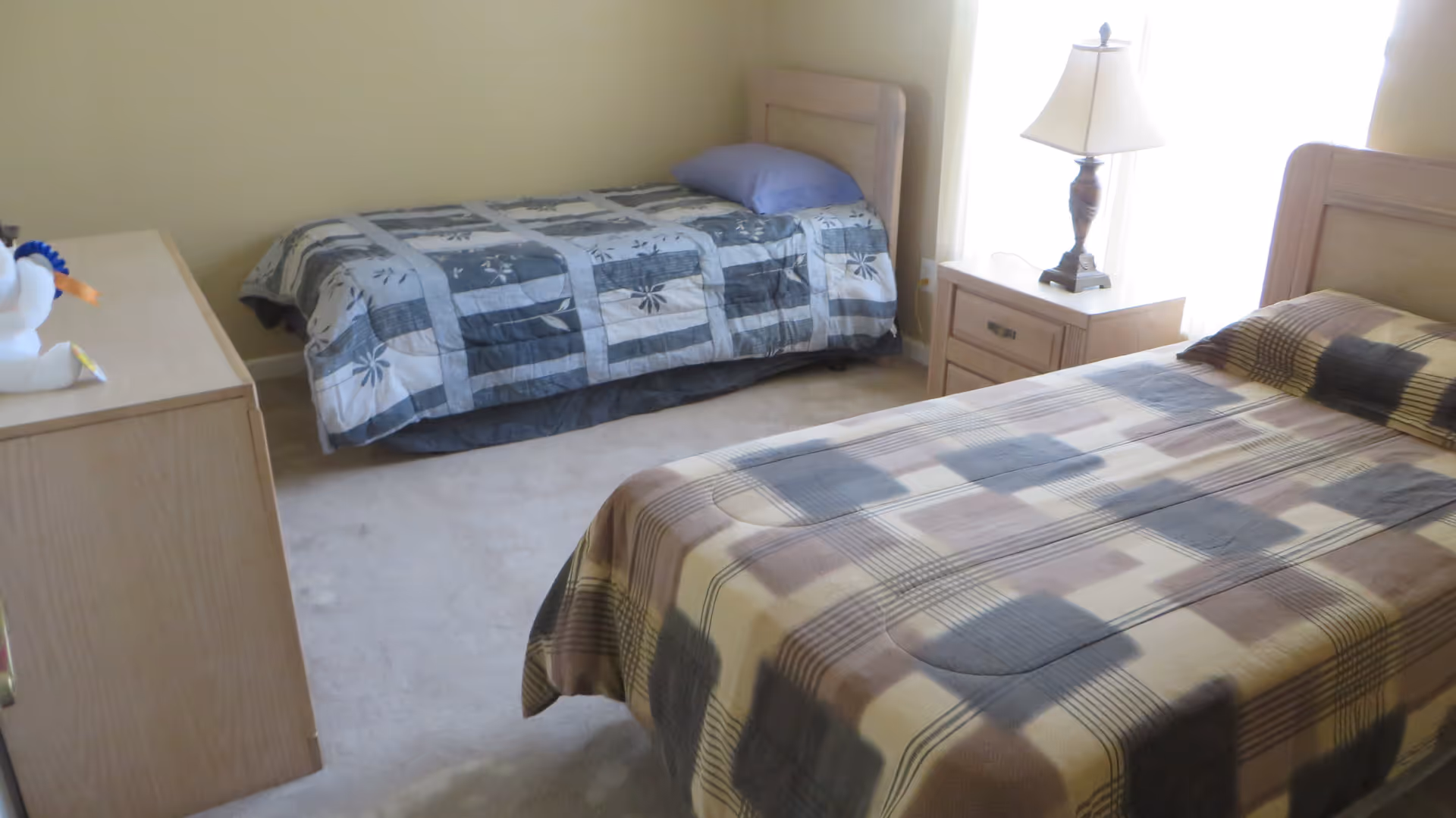 Small bedroom with two twin beds, a nightstand and lamp, and a dresser with a stuffed toy.