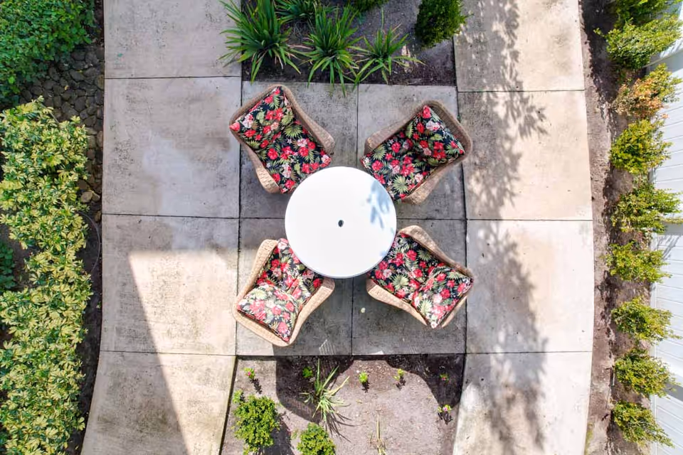 Aerial view of an outdoor patio area with a round white table surrounded by four wicker chairs with floral cushions, set on a concrete tiled surface with garden beds and green plants around the edges.