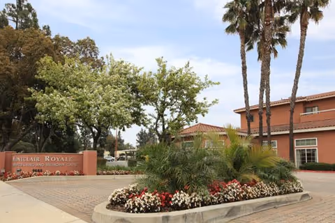 Entrance area of Montclair Royale, a memory care facility, featuring a landscaped roundabout with palm trees, flowering plants, and a sign displaying the facility's name. The building is visible in the background under a partly cloudy sky.