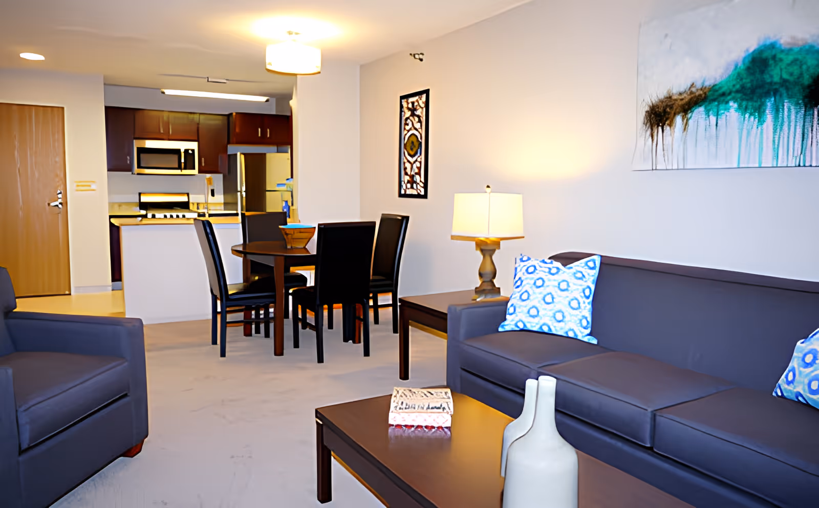 A modern living area with a dark gray sofa adorned with blue and white patterned pillows, a matching armchair, a wooden coffee table with decorative items, and a dining table with four chairs. The background shows a kitchen area with wooden cabinets, a microwave, and a refrigerator. The room is well-lit with ceiling and table lamps, and there is abstract artwork on the walls.