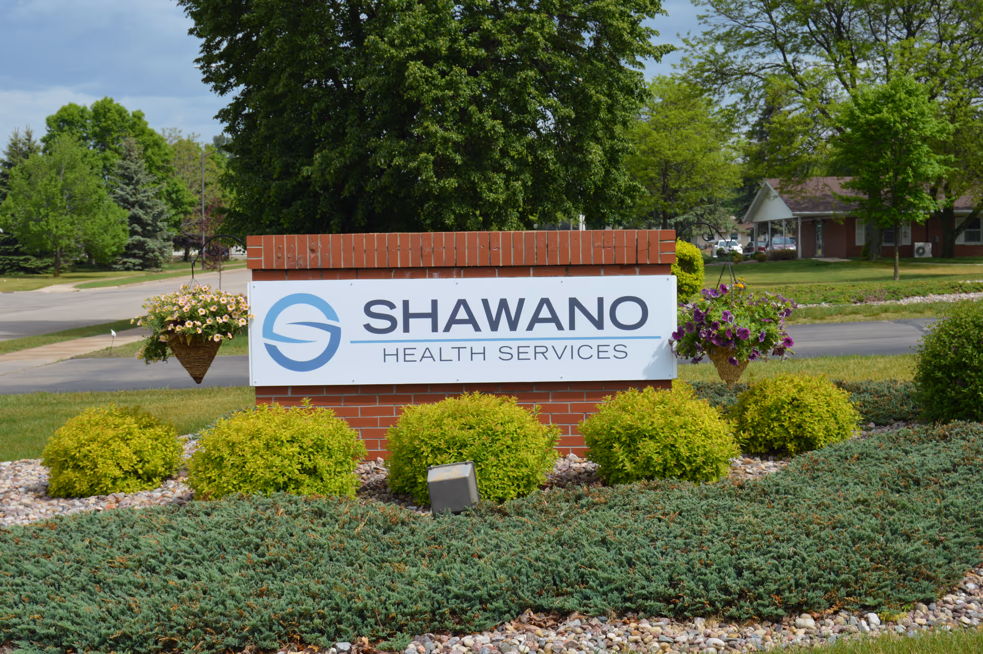 A brick sign with a white panel displaying the logo and text 'Shawano Health Services' surrounded by green bushes and hanging flower baskets, with trees and a road in the background.