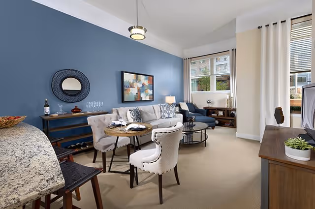 A bright and modern living room with a blue accent wall, a round mirror, and abstract artwork. The room features a beige sofa with patterned cushions, a round wooden dining table with four upholstered chairs, a glass coffee table, and a wooden TV stand with a flat-screen TV. Large windows with white curtains allow natural light to fill the space.
