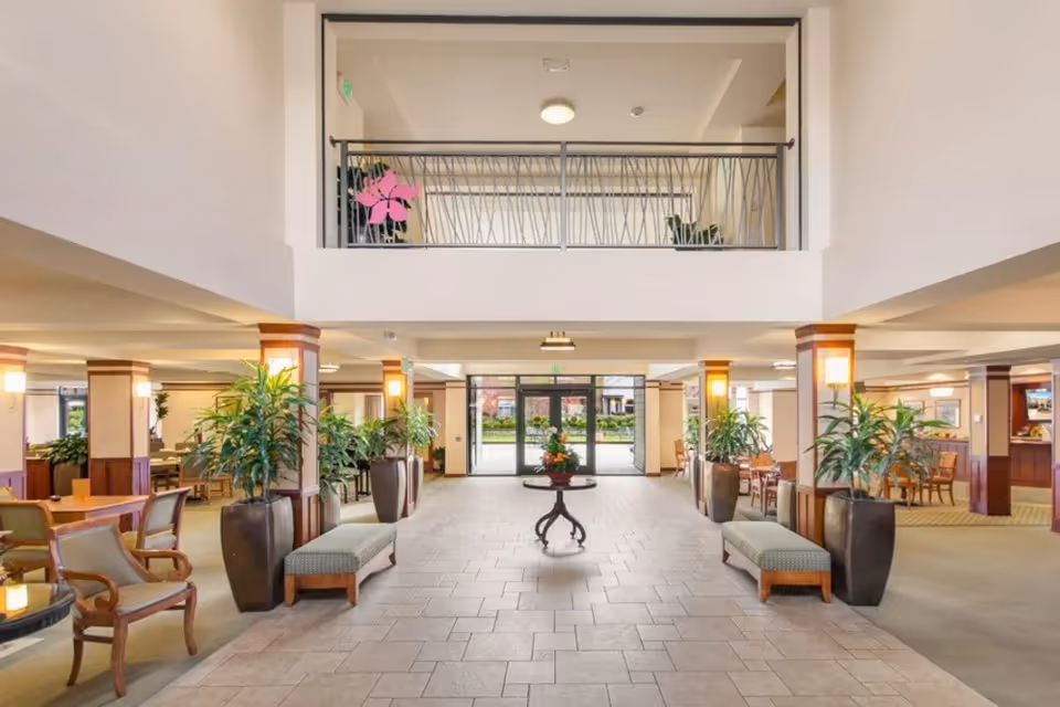 Spacious and well-lit interior common area of Merrill Gardens at The University featuring tiled flooring, seating benches, potted plants, wooden chairs and tables, and a central round table with a floral arrangement. Large glass doors lead outside, and there is a second-floor balcony with a decorative railing.