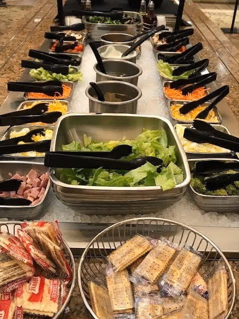 A salad bar with various fresh vegetables and toppings including lettuce, broccoli, shredded carrots, celery, diced ham, and other salad ingredients in metal containers with black serving tongs. In front of the salad bar, there are baskets filled with individually wrapped crackers.