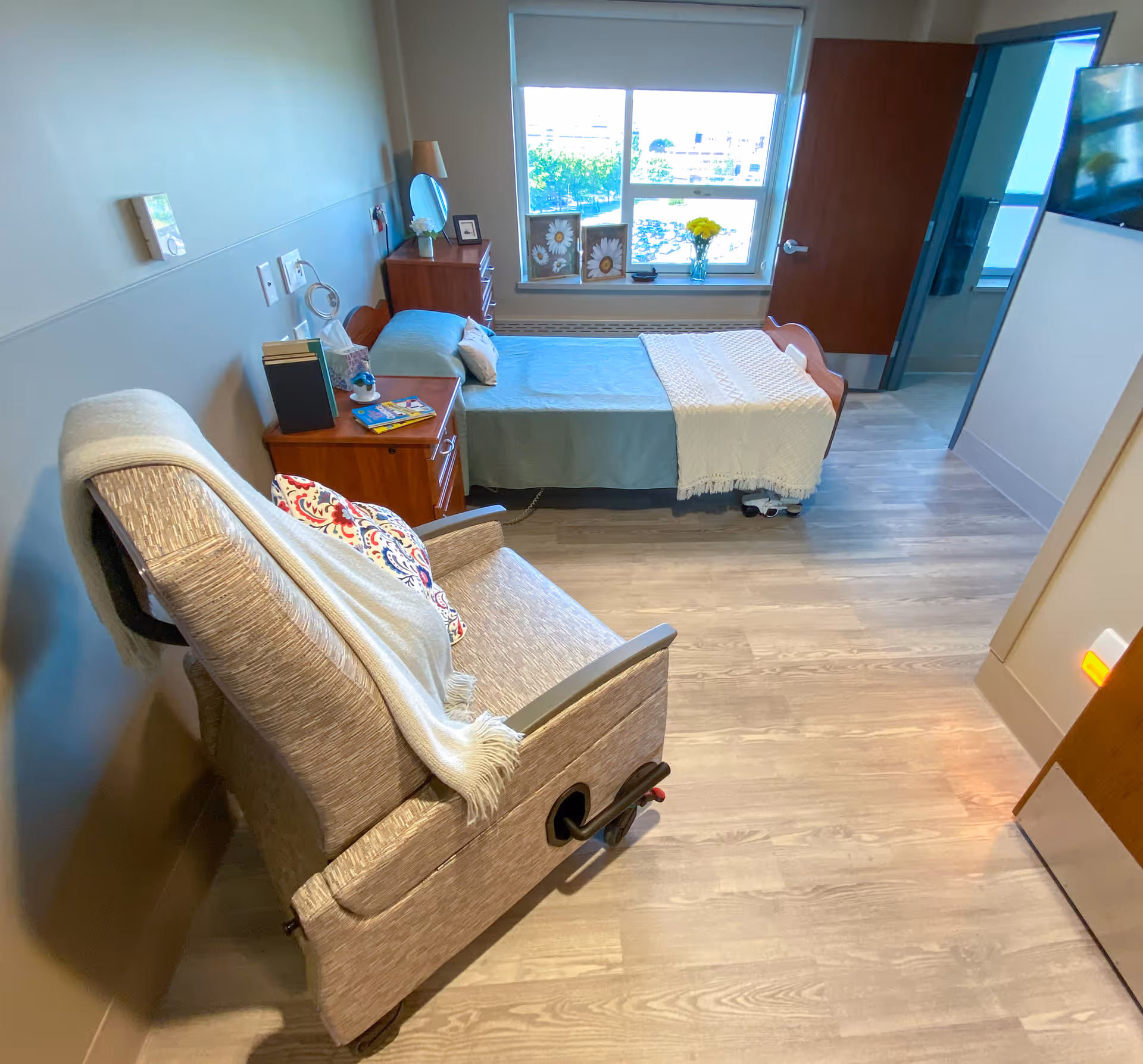 A tidy senior living bedroom with a single bed, bedside table, recliner chair, window, and wall-mounted TV.