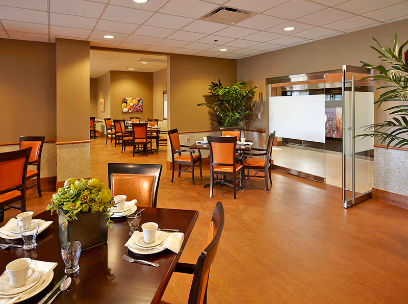 A spacious dining room with several wooden tables and chairs arranged neatly. Each table is set with white plates, cups, napkins, and silverware. There are green plants placed around the room, and a glass door with frosted panels is visible on the right side. The walls are painted in warm beige tones, and there is a painting of fruit on the far wall.