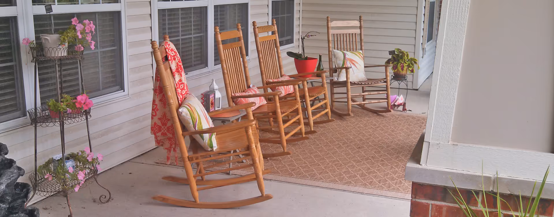 Front porch with several wooden rocking chairs, cushions and potted plants set beside a building.