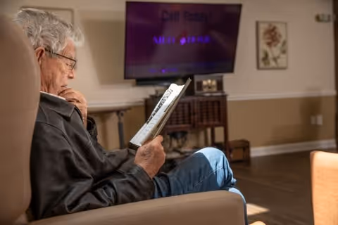 An elderly man with gray hair and glasses is sitting in a comfortable chair, reading a book in a cozy living room. In the background, there is a television mounted on the wall and a small wooden table with decorative items.