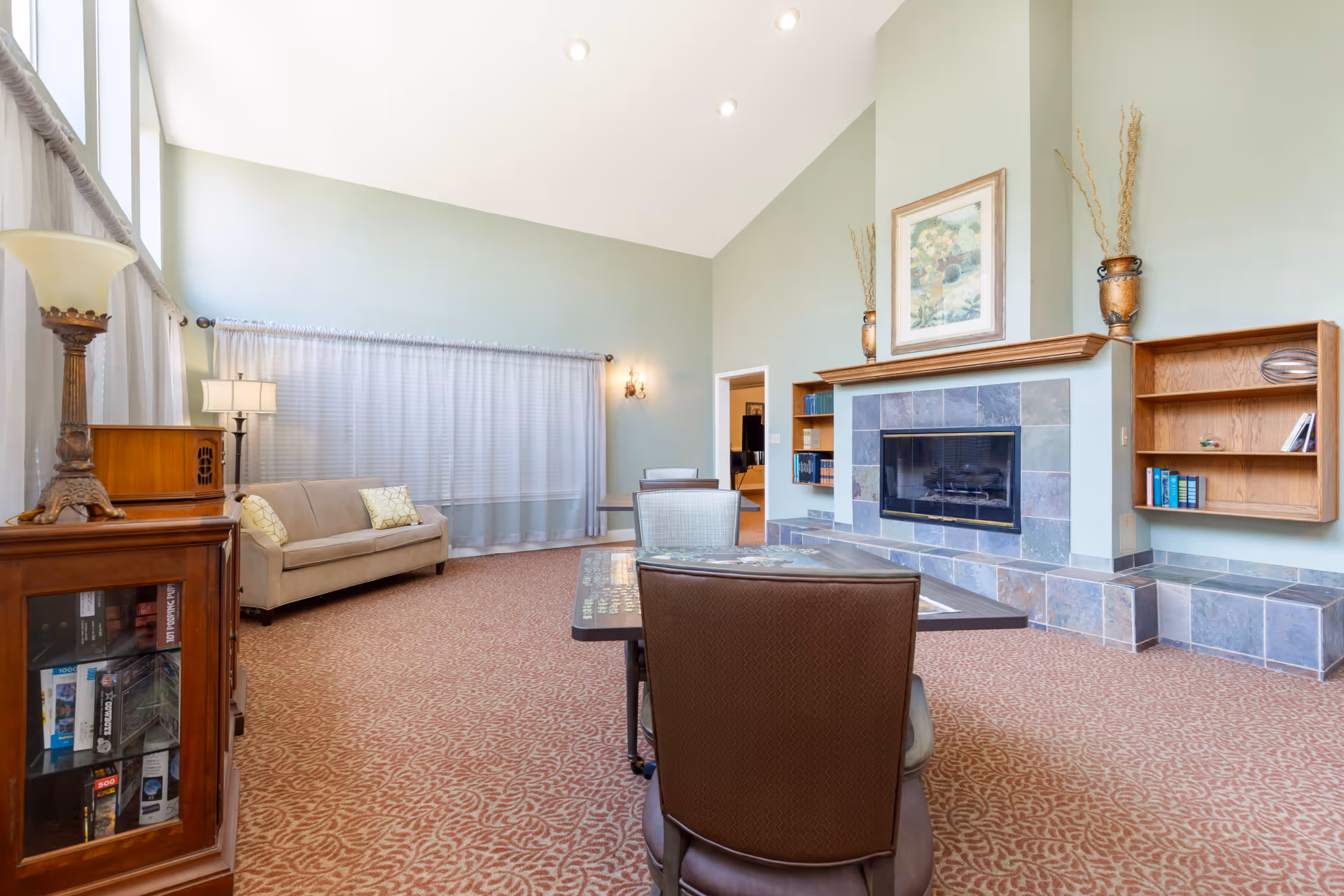 A spacious living room with high ceilings painted in light green. The room features a beige sofa with two patterned cushions, a wooden cabinet with a lamp, a fireplace with a tiled surround and a wooden mantel decorated with vases and a framed painting. There are built-in wooden shelves with books on the right side of the fireplace. A table with chairs is positioned in the foreground on a patterned carpeted floor. Large windows with sheer curtains allow natural light into the room.