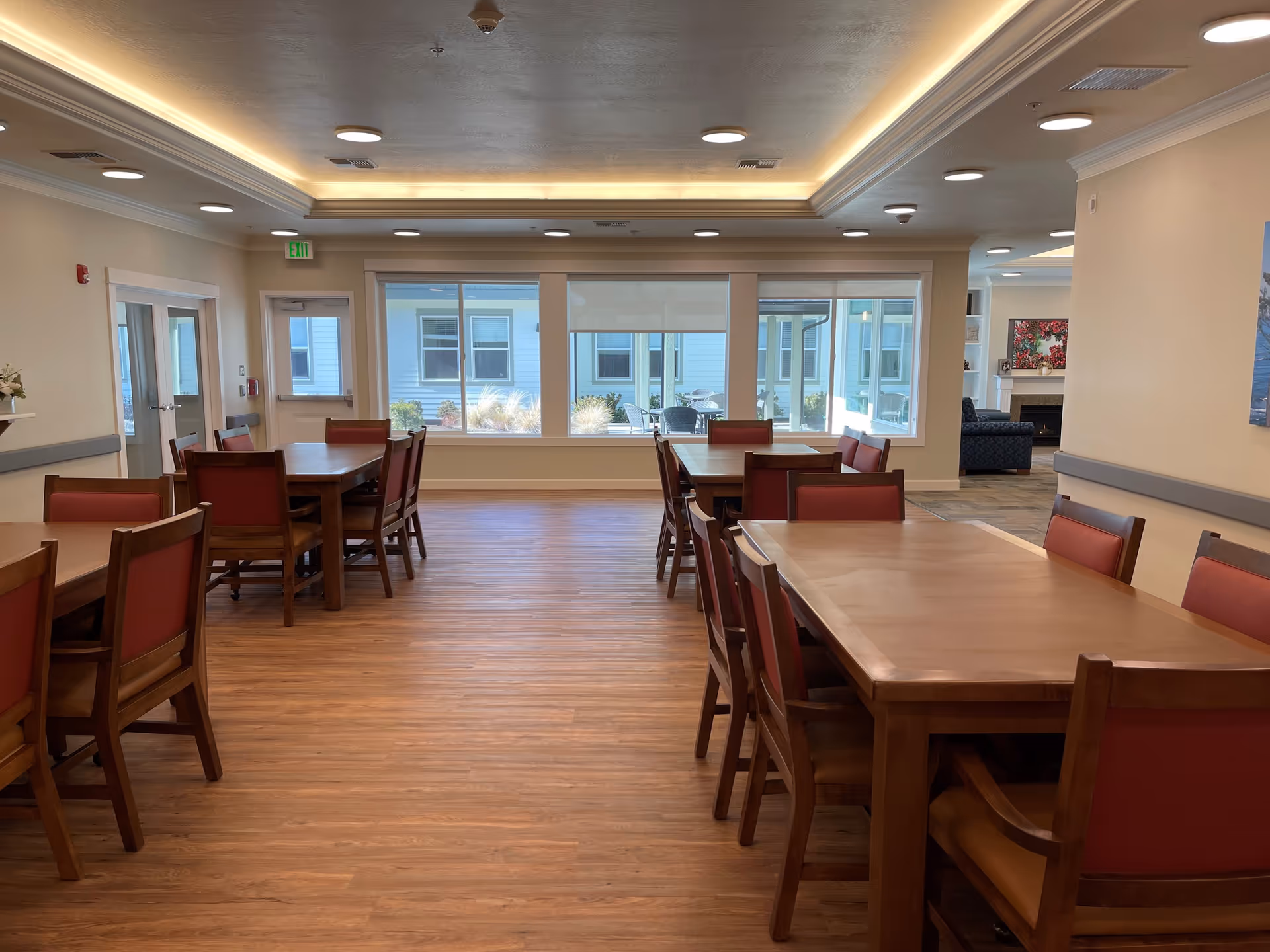 A bright dining room with wooden tables and chairs featuring red cushions. Large windows provide natural light and a view of an outdoor patio area. The room has a wooden floor and recessed ceiling lights with a modern ceiling design.