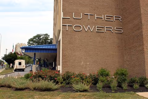 Exterior view of a brick building with the sign 'Luther Towers' mounted on the wall. There is a covered entrance with a blue roof and some bushes and greenery in front of the building. A vehicle is parked near the entrance and other buildings are visible in the background.