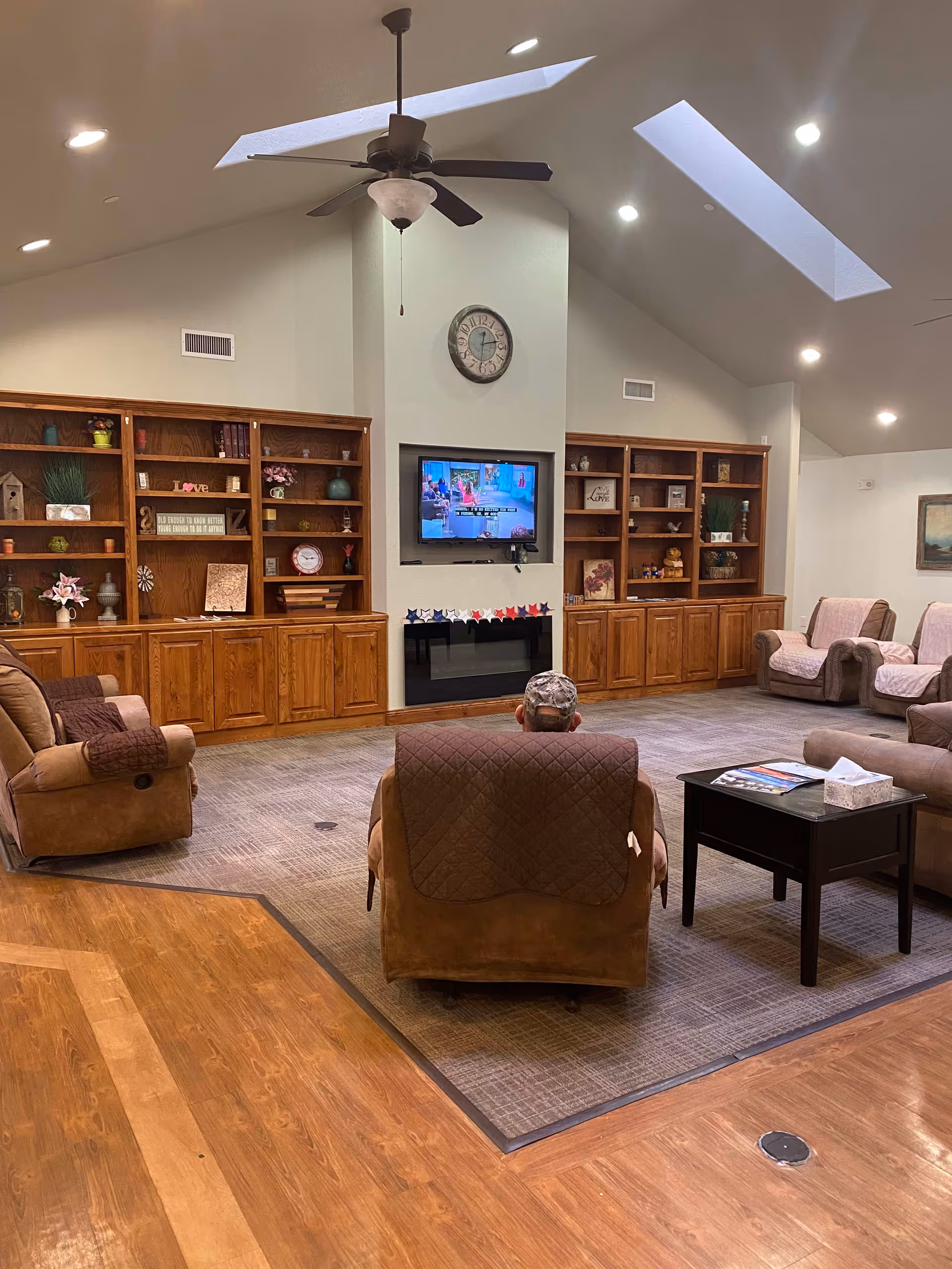 A cozy senior living room with a high vaulted ceiling and skylights. The room features wooden built-in shelves filled with decorative items, a wall-mounted TV above an electric fireplace, and several comfortable armchairs arranged around a central coffee table. A person wearing a cap is seated in one of the armchairs facing the TV. The floor is a combination of wood and carpet.