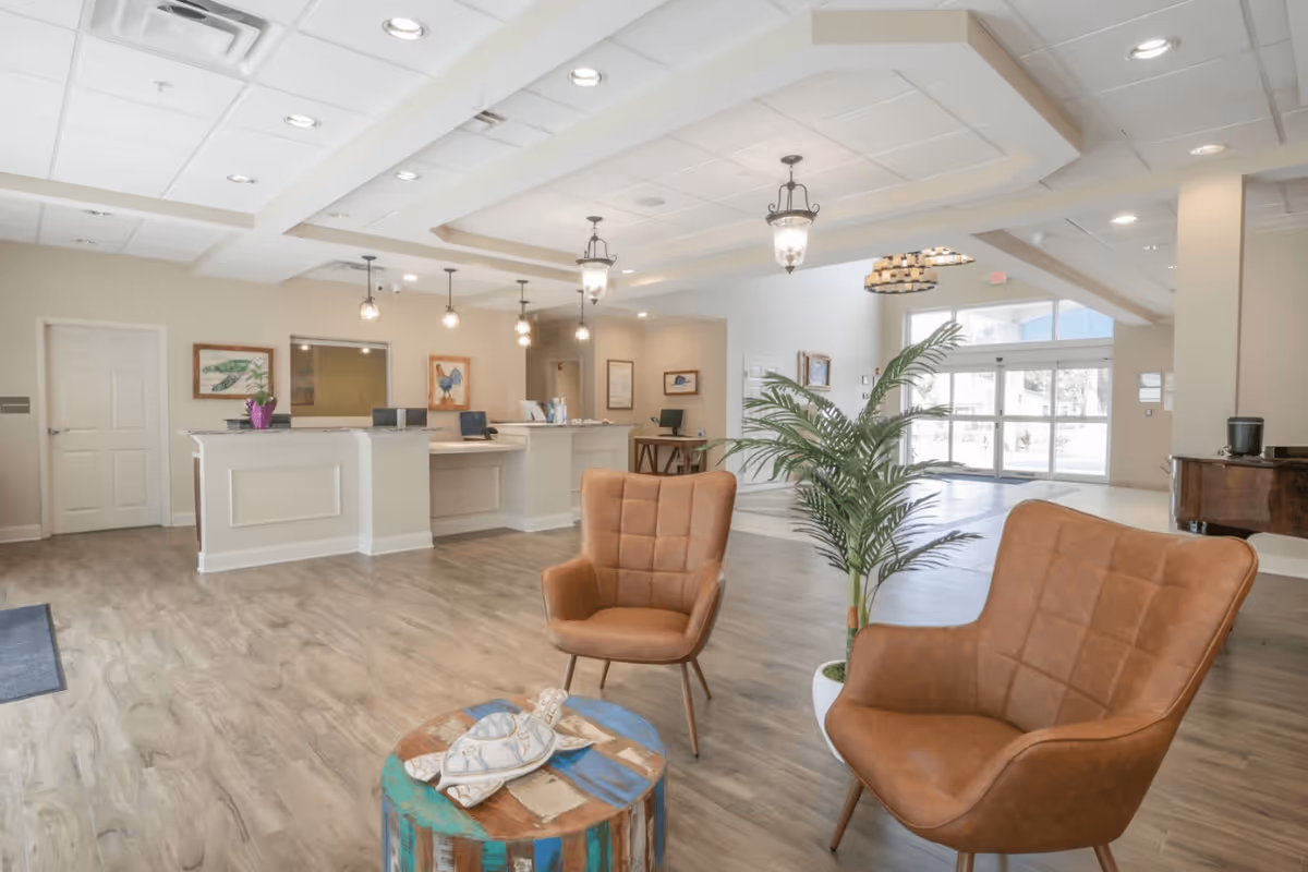 A spacious lobby reception area featuring two brown armchairs, a wooden table with decorative items, and a welcoming front desk with computers. Natural light streams in through large windows, enhancing the inviting atmosphere.