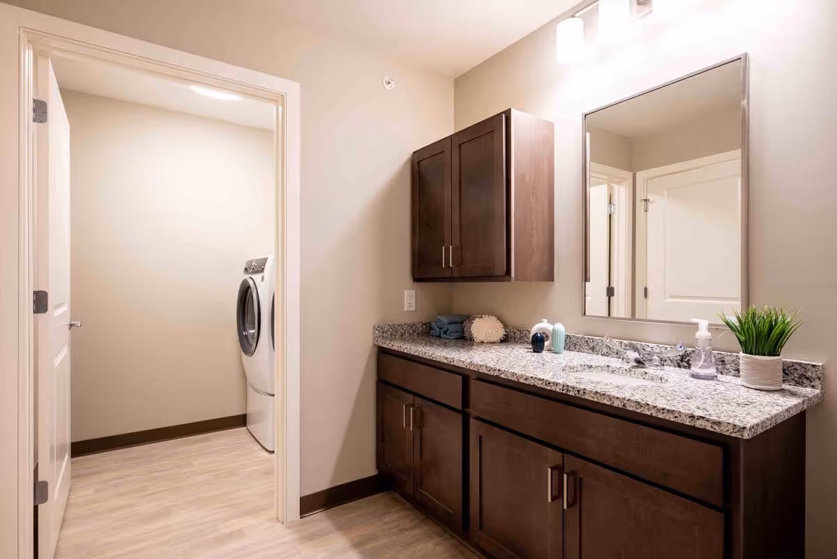 Bathroom with dark wood vanity and granite countertop, mirror above the sink, and an adjacent laundry alcove with a front-loading washer.