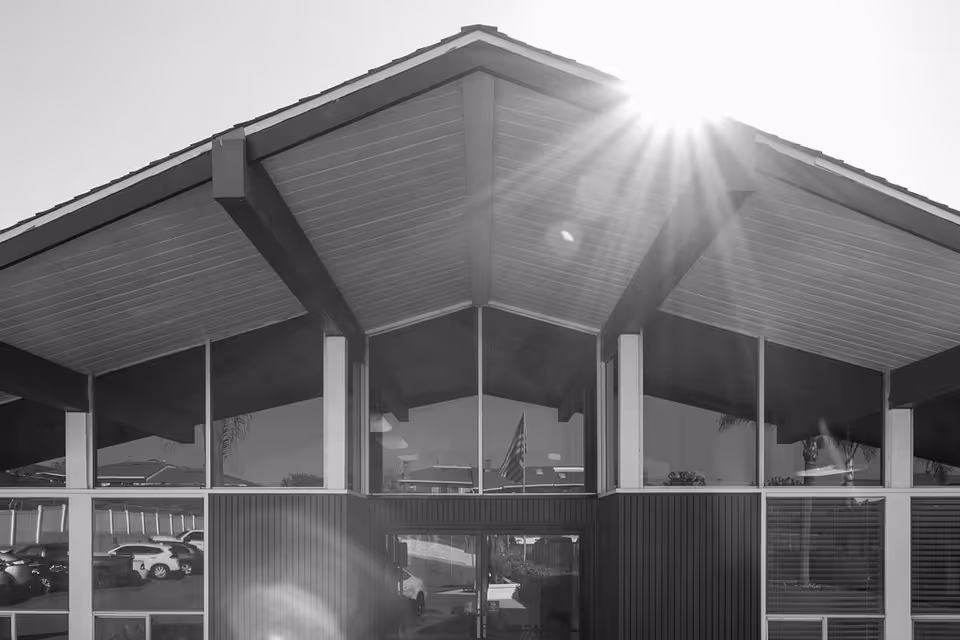Black and white photo of the front exterior of a building with a peaked roof and large windows reflecting parked cars and an American flag. Sunlight is shining from the top right corner.