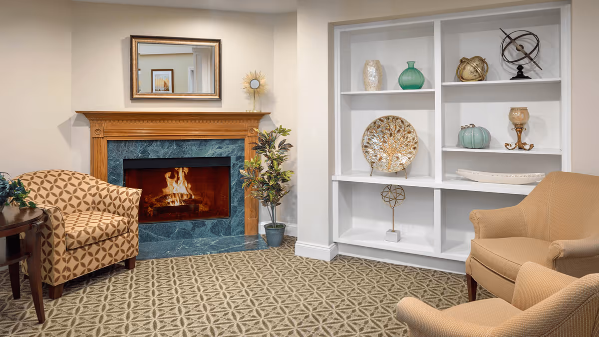 A cozy living room area with a lit fireplace surrounded by green marble and a wooden mantel. Above the fireplace is a rectangular mirror. To the left is a patterned armchair next to a small wooden table with a plant. To the right are two beige armchairs and a white built-in shelving unit displaying decorative items including vases, a decorative plate, and sculptures. The floor is carpeted with a geometric pattern.