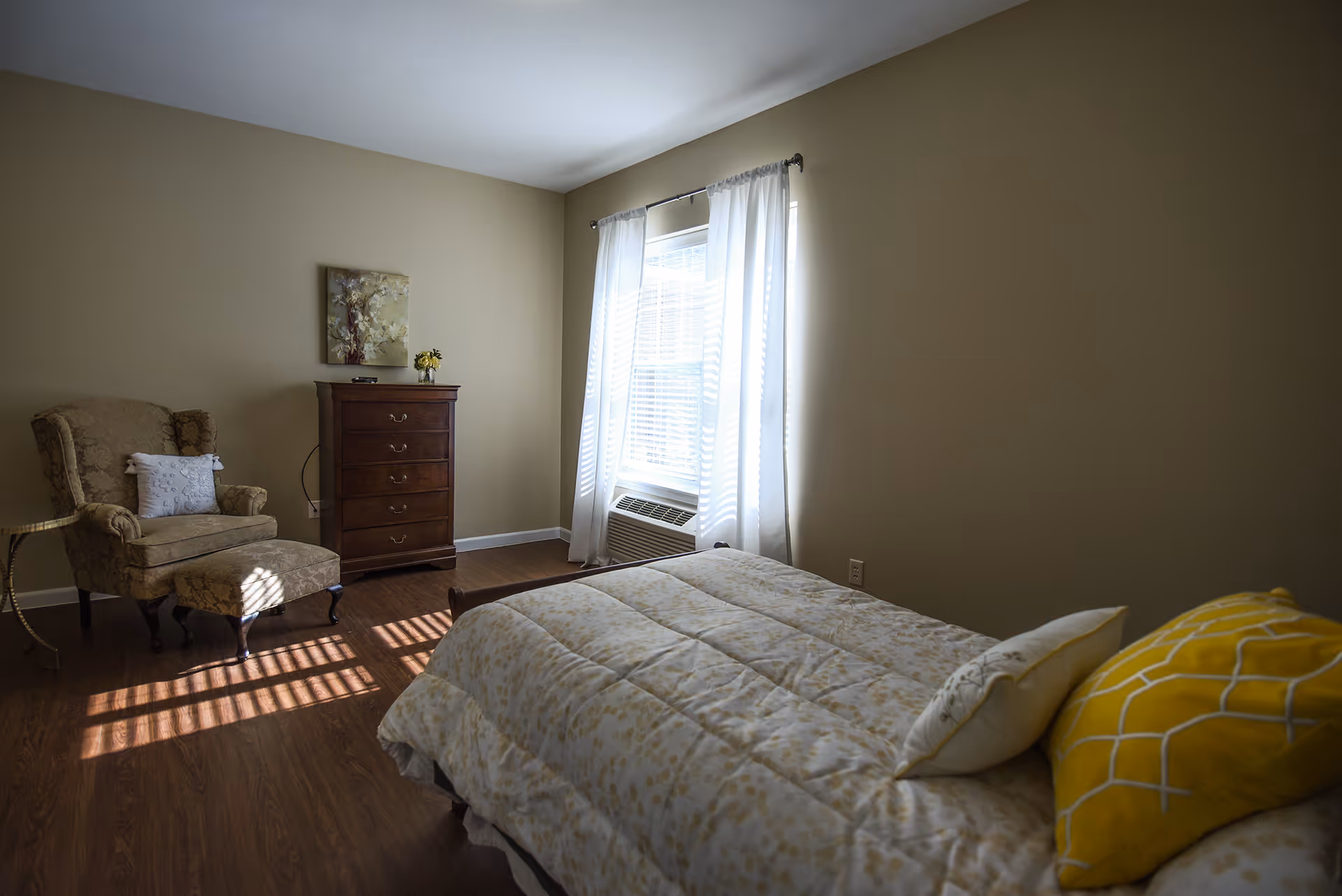 A bedroom with a bed covered in a light-colored comforter and two pillows, one yellow and one white. There is a window with white curtains letting in sunlight, a wooden chest of drawers with a small vase of flowers on top, and an upholstered armchair with a matching ottoman. The walls are beige and the floor is wood.