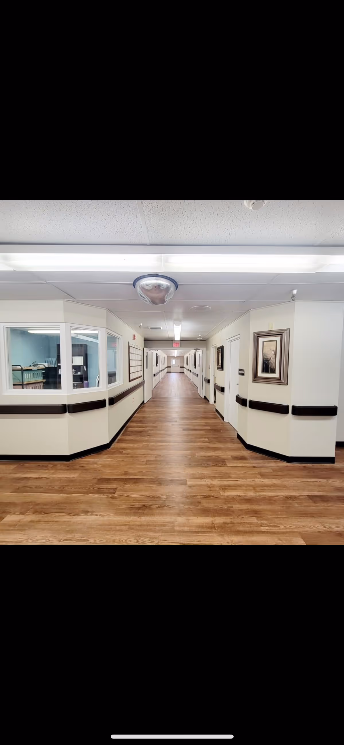 A long, clean hallway in a skilled nursing facility with wood-patterned flooring, white walls, handrails along both sides, framed artwork on the walls, and a nurse's station with windows on the left side.
