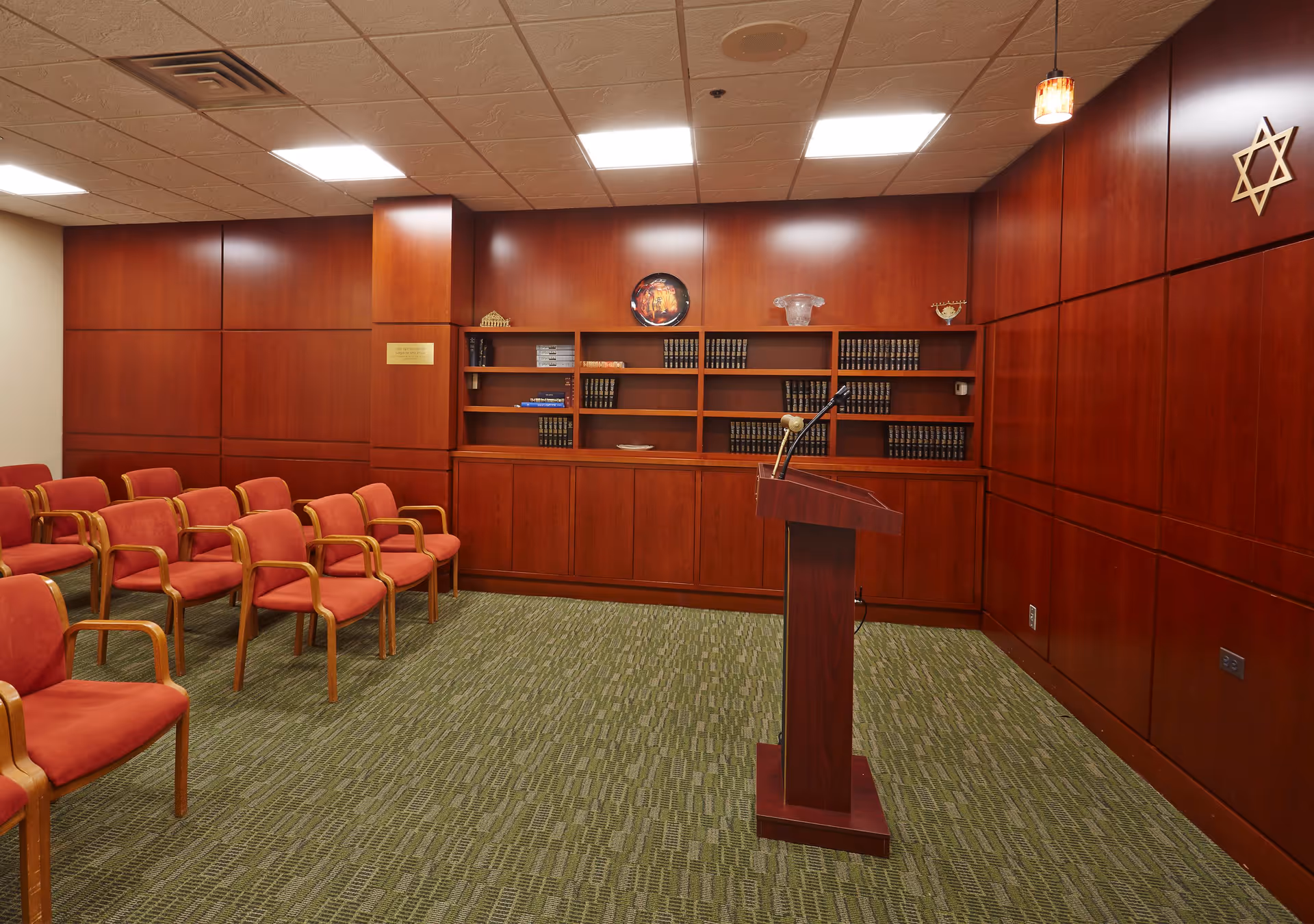A small meeting or lecture room with red cushioned chairs arranged in rows facing a wooden podium with a microphone. The room has wood-paneled walls, built-in shelves with books and decorative items, a green patterned carpet, and a Star of David symbol mounted on the wall.