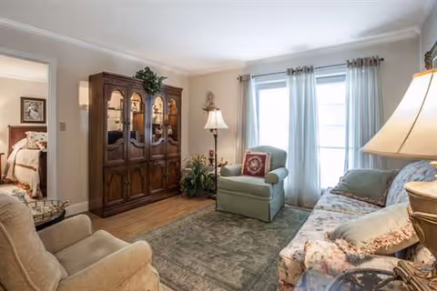 A cozy living room with a floral patterned sofa, a green armchair with a decorative pillow, a beige recliner, a wooden cabinet with glass doors, a floor lamp, and large windows with sheer curtains allowing natural light to fill the room. A doorway leads to a bedroom with a bed visible.