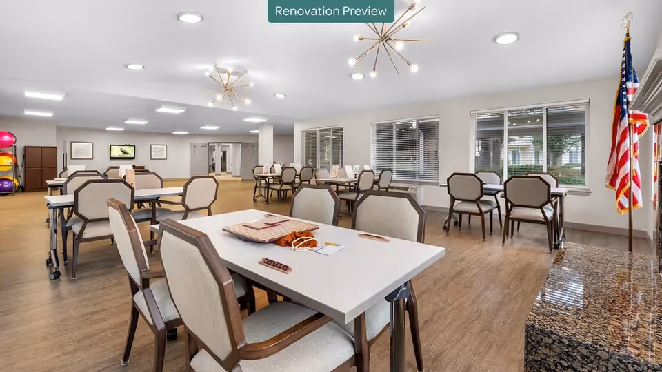 A spacious, well-lit common area with multiple tables and chairs arranged for group activities or dining. The room has large windows with blinds, modern light fixtures on the ceiling, and an American flag displayed near a granite countertop. There are some colorful exercise balls stacked in the back corner and framed pictures on the far wall. A sign at the top center reads 'Renovation Preview'.