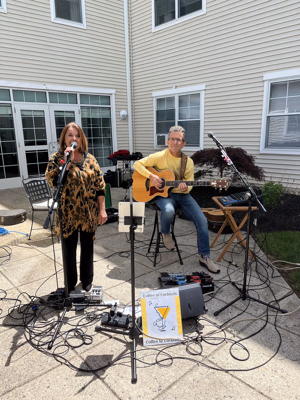 A woman singing into a microphone and a man playing an acoustic guitar while seated on a stool, performing outdoors on a paved patio area in front of a beige building with white-framed windows and doors. Various musical equipment and cables are visible on the ground, along with a sign that reads 'Coffee to Cocktails.'