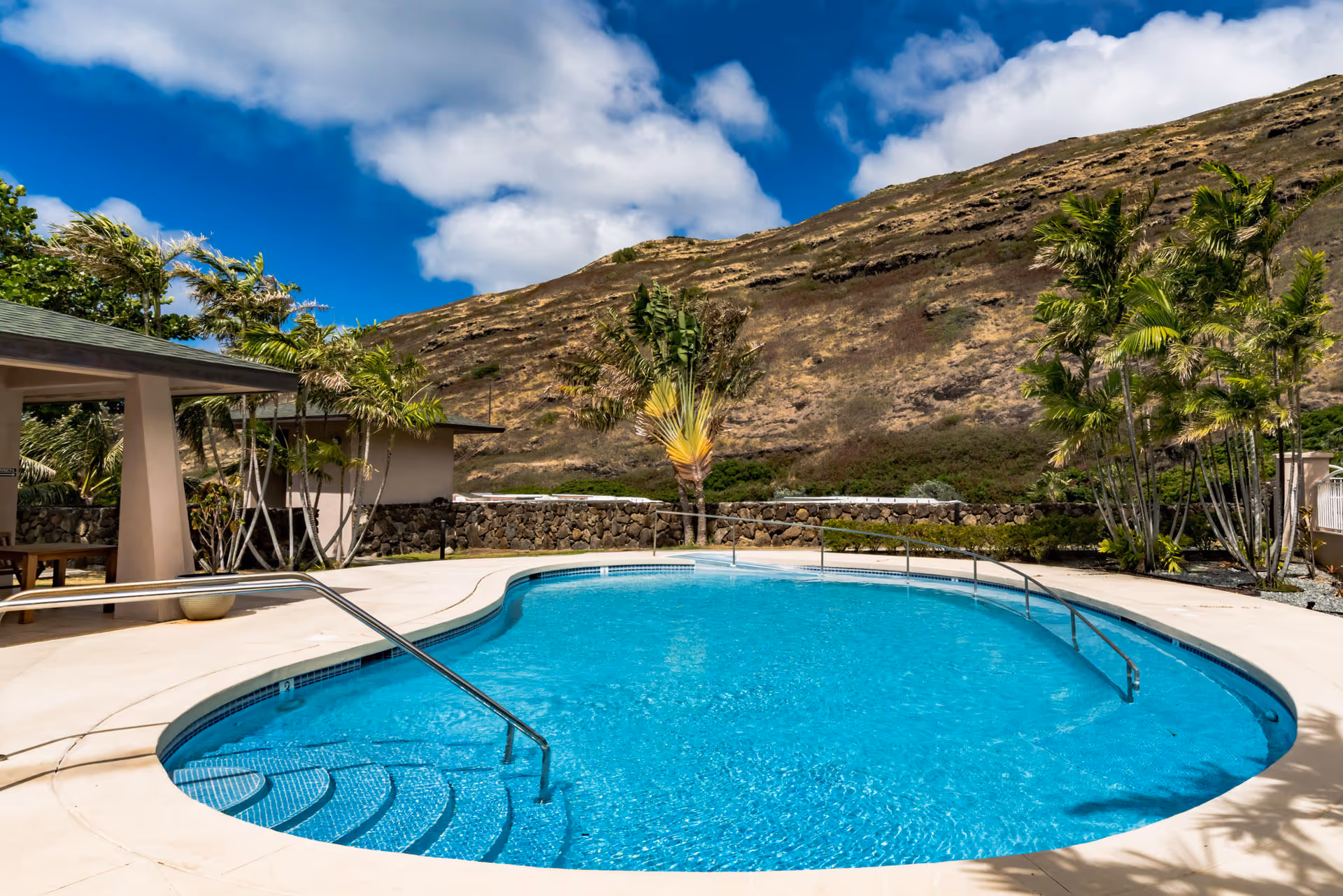 Outdoor swimming pool with clear blue water surrounded by a concrete deck, palm trees, and a small covered seating area. In the background, there is a rocky hillside under a partly cloudy blue sky.