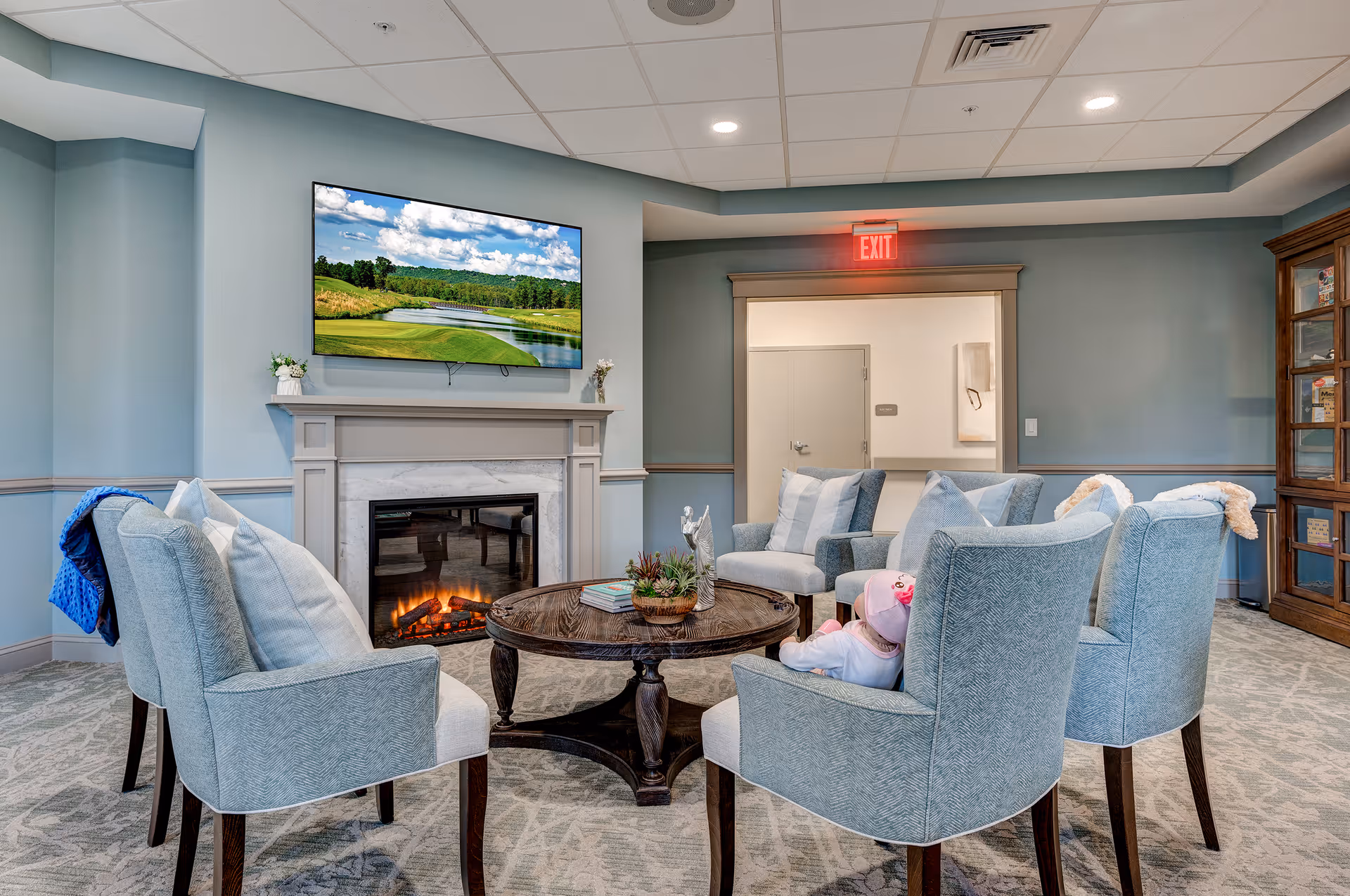 A cozy sitting area with six light blue upholstered chairs arranged around a round wooden coffee table. The room features a fireplace with a TV mounted above it displaying a scenic golf course. The walls are painted light blue with white trim, and there is an exit sign above a doorway in the background. A wooden cabinet with glass doors is visible on the right side of the room.
