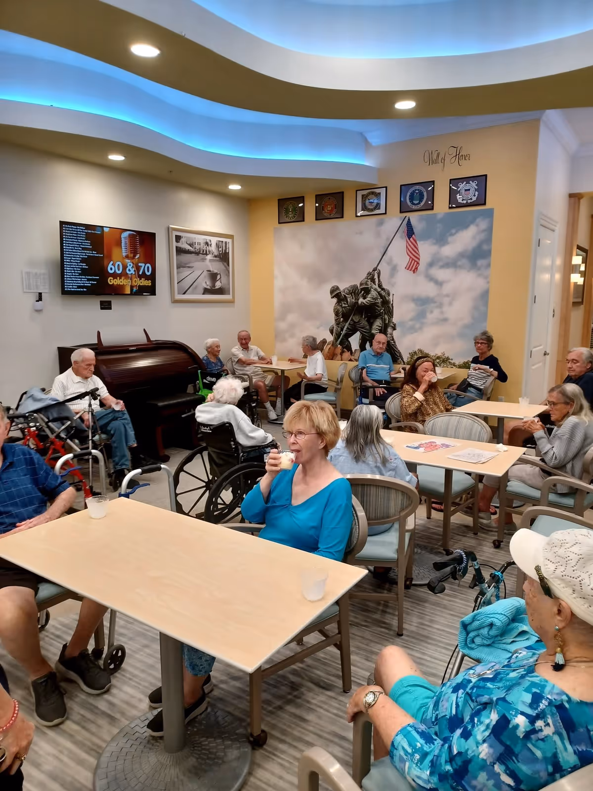 A group of elderly people sitting around tables in a common room of an assisted living facility. Some are in wheelchairs, and others are seated in chairs. The room has a ceiling with blue accent lighting, a piano, a wall-mounted TV displaying '60 & 70 Golden Oldies,' and a large mural of the Iwo Jima flag raising with the American flag. The wall above the mural reads 'Wall of Honor' with military emblems displayed.