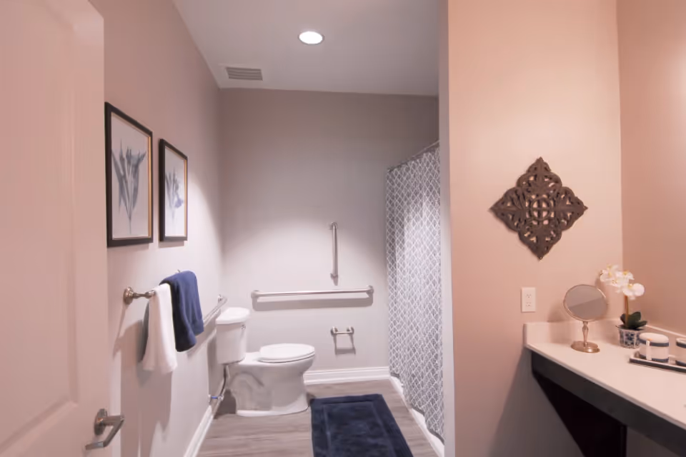 A clean and modern bathroom with a toilet, a shower with a patterned curtain, a towel rack holding white and navy blue towels, two framed botanical prints on the wall, a dark blue bath mat on the floor, and a vanity area with a small round mirror, decorative plant, and toiletries.
