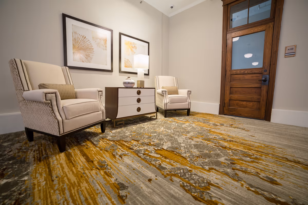 A small seating area with two beige upholstered armchairs separated by a modern three-drawer side table with a lamp and decorative vase on top. Two framed abstract artworks hang on the wall above the table. The floor is covered with a patterned carpet in shades of gray and mustard yellow. A wooden door with glass panels is visible on the right side of the image.