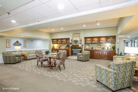 A spacious senior living facility common area with patterned carpet, multiple upholstered chairs and sofas arranged around small tables, a fireplace with a mirror above it, and wooden cabinetry with glass doors displaying decorative items. The room is well-lit with ceiling lights and lamps, and there are plants and framed artwork on the walls.