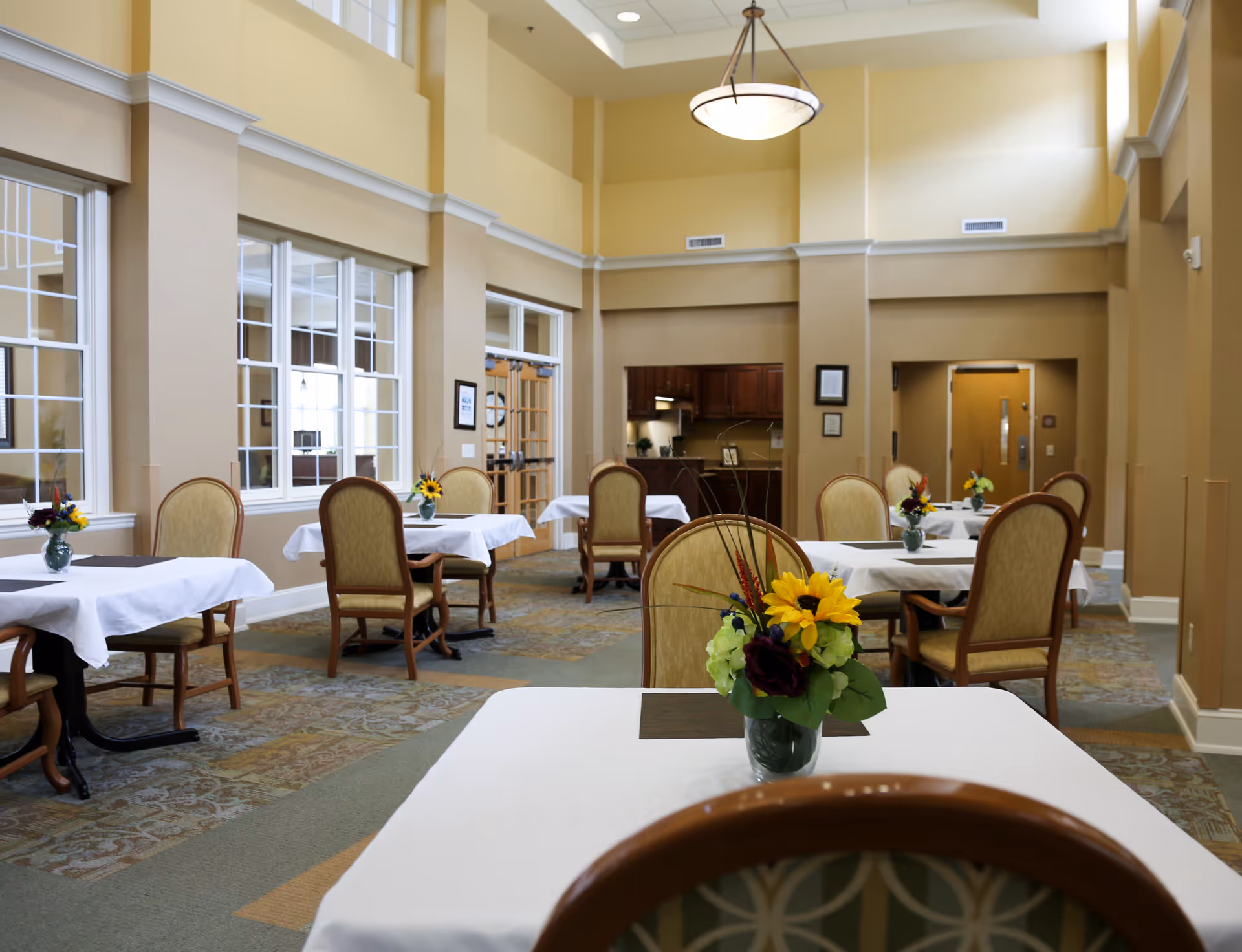A bright dining room with several tables covered in white tablecloths, each adorned with a small vase of colorful flowers. The room has high ceilings, large windows, beige walls, and carpeted floors. Wooden chairs with cushioned seats surround the tables. In the background, there is a small kitchen area with dark wooden cabinets.