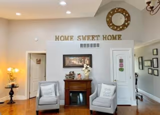 A cozy living room area with two gray armchairs placed on either side of a wooden electric fireplace. Above the fireplace is a framed picture and a vase with flowers. On the wall above, gold letters spell out 'HOME SWEET HOME'. To the left, a small round table with a lamp and decorative items is visible. The room has light-colored walls, wooden flooring, and recessed ceiling lights.